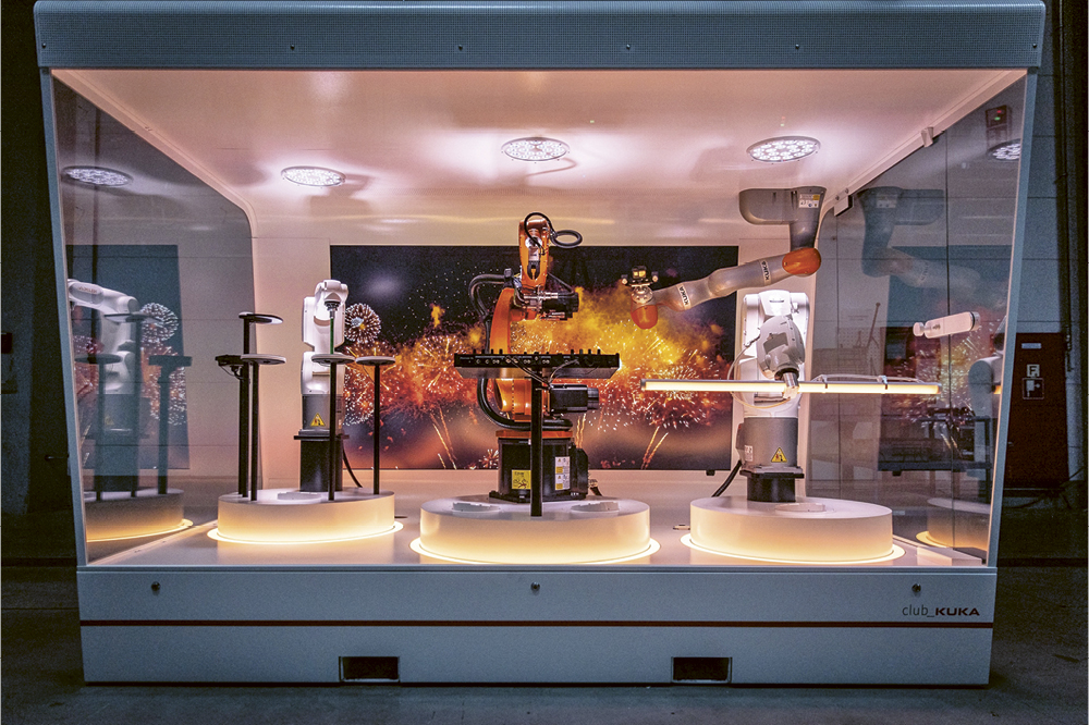 Robots play drums and keyboard in a glass room on a show stage with an image of fireworks in the background