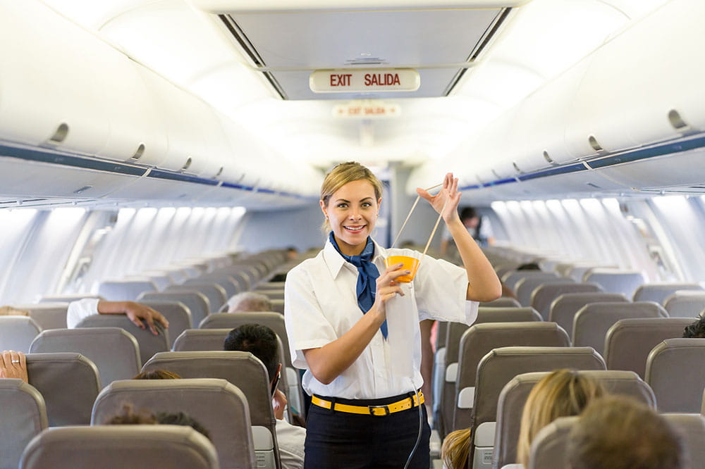 Smiling female flight attendant demonstrating an airline safety oxygen mask inside a commercial airplane cabin,