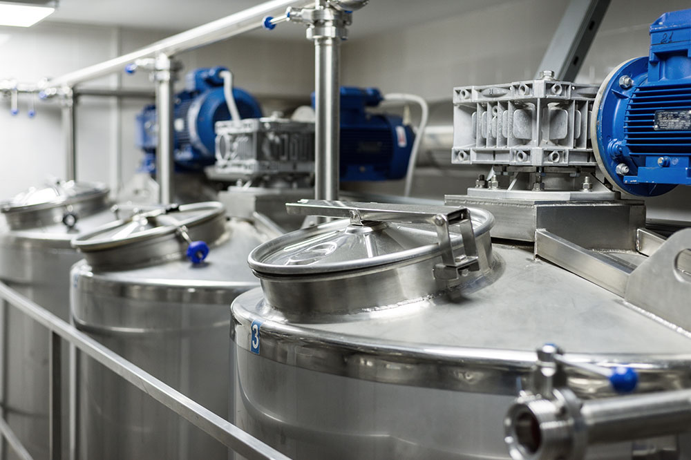 Row of large stainless steel industrial tanks with closed lids and blue electric motors in a processing facility.