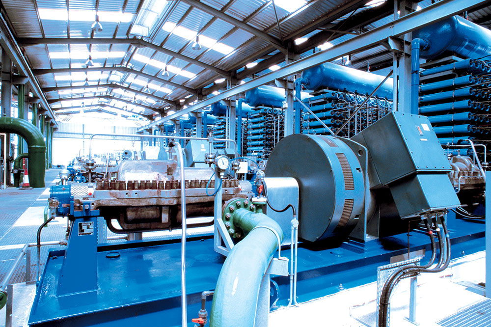 Interior view of an industrial power plant with generator and turbine engines, high-performance pump system, steel pipes, pressure gauges, and heat exchanger units in a modern factory hall with metal roof construction. 