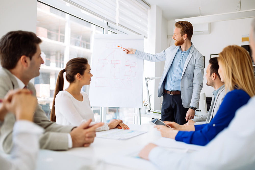 Male in business suit points on a drawing on a flip chart while several people are listening
