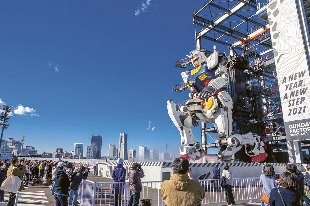 Haushoher Roboter kniet auf Plattform mit Skyline im Hintergrund und Menschenmenge, die ihn fotografiert.  Copyright: Dick Thomas Johnson 