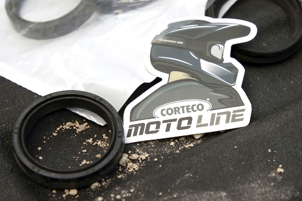 Ein Sticker von einer Person mit einem Motorradhelm auf dem Kopf mit der Aufschrift Corteco Moto Line liegt auf einem Tisch neben einer Dichtung. 