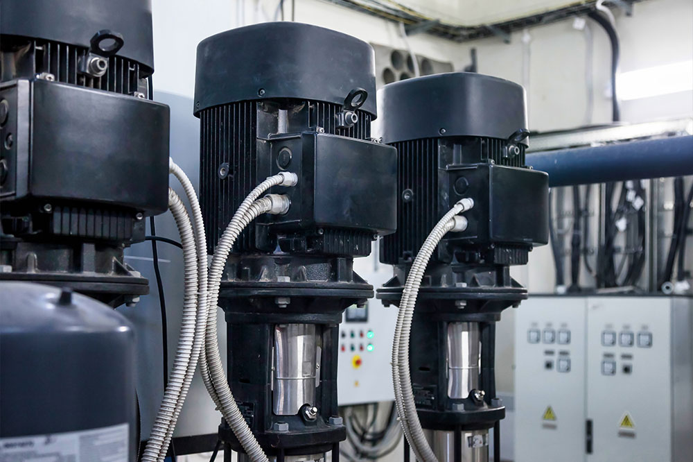 Innenansicht eines Raumes mit drei Wasserpumpen, Rohren und elektrischen Leitungen.