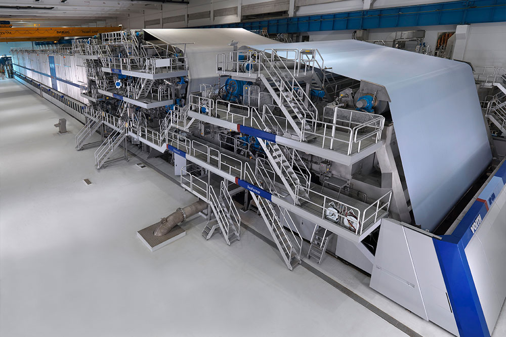 Industrielle Papierherstellungsmaschine in einer modernen Fabrikhalle, in der eine Endlosrolle aus weißem Papier produziert wird, mit Metalltreppen, Sicherheitsgeländern, Steuerungsplattformen und schwerem Industriegerät
